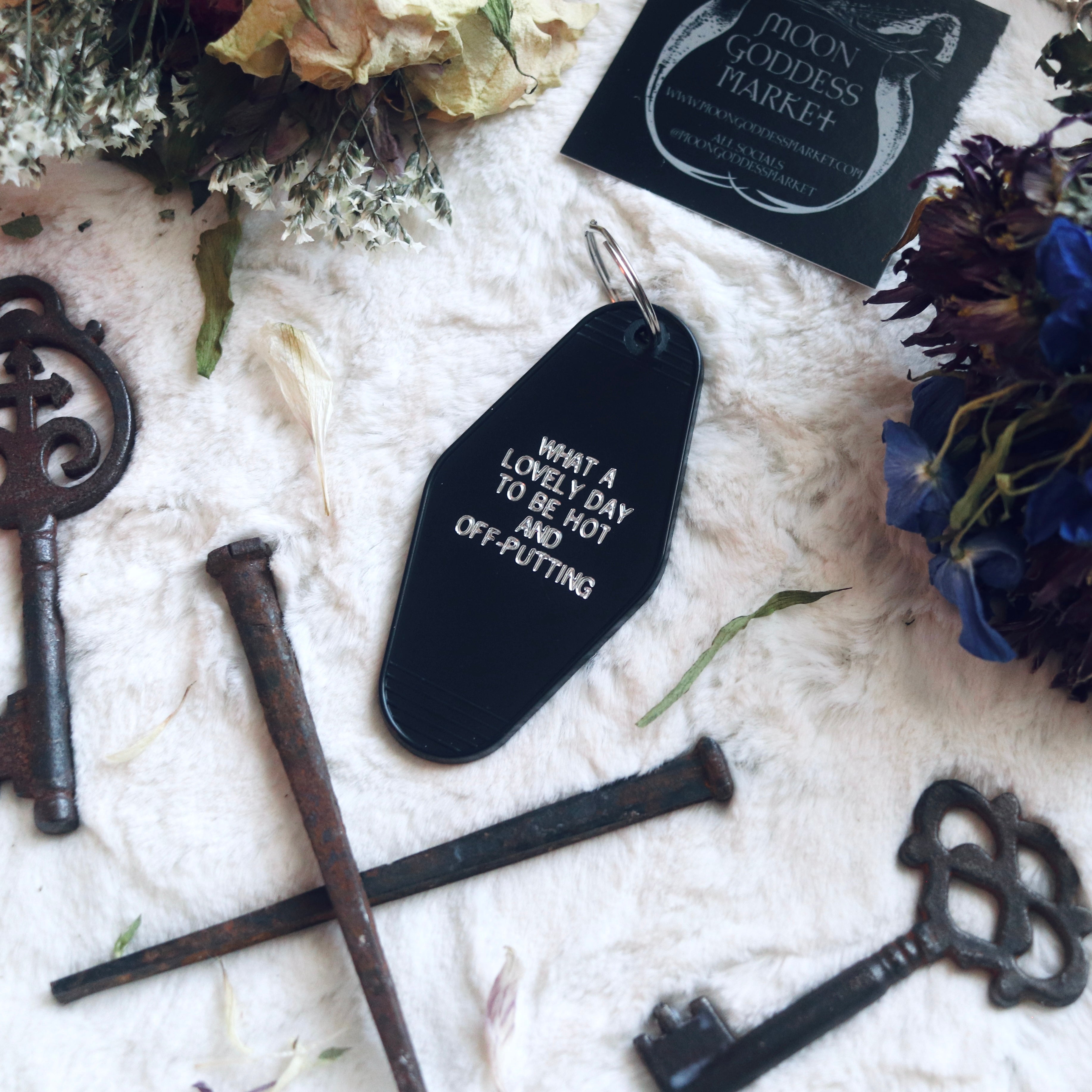 A black keychain with silver text 'WHEN A LOVELY DAY TO BE HOT AND OFF-PUTTING' printed on it, displayed along with a decorative floral arrangement and vintage keys.